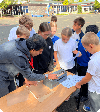A group of young primary school children engaged in a tactile learning activity with a Braille writing machine